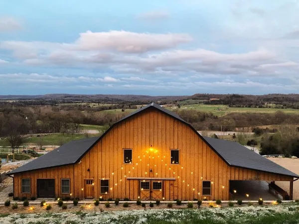 A large wooden barn with string lights and a metal roof set in a rural landscape with rolling hills in the background.