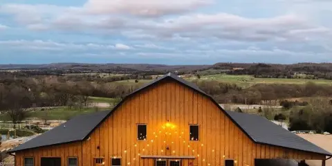 A large wooden barn with string lights and a metal roof set in a rural landscape with rolling hills in the background.