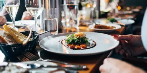 A close-up of a plated gourmet dish with a garnish and sauce, served at a table with wine glasses and a bread basket.