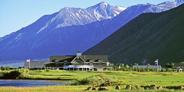 A large clubhouse building with a dark roof and white trim sits on a manicured green lawn, backed by a large mountain range with snow-capped peaks.