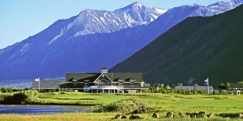 A large clubhouse building with a dark roof and white trim sits on a manicured green lawn, backed by a large mountain range with snow-capped peaks.