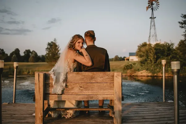 A bride and groom sitting on a rustic wooden bench by a pond with a windmill in the background.
