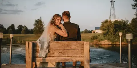A bride and groom sitting on a rustic wooden bench by a pond with a windmill in the background.