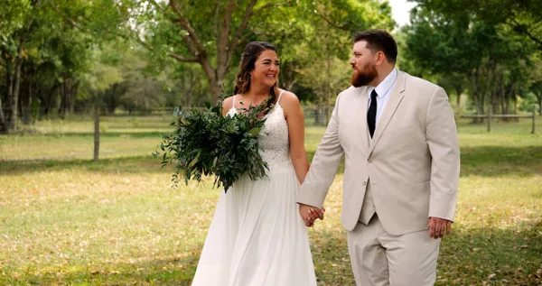 A bride holding a green bouquet and a groom in a beige suit walking hand-in-hand in a grassy field with trees in the background.