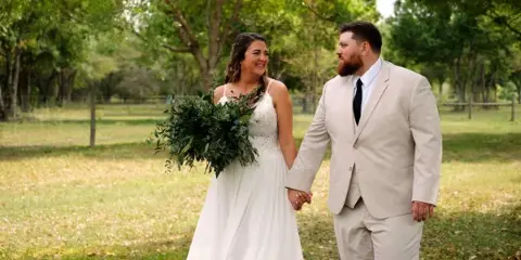 A bride holding a green bouquet and a groom in a beige suit walking hand-in-hand in a grassy field with trees in the background.
