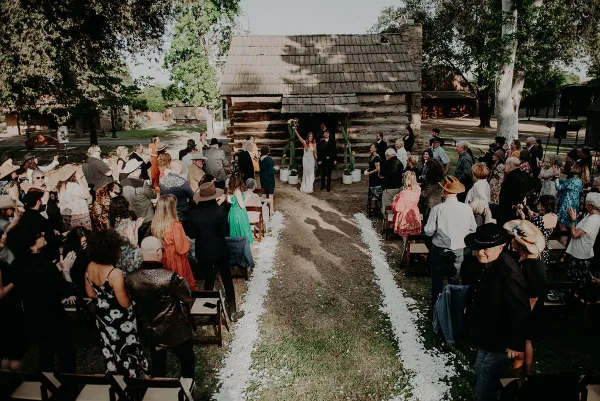 A wedding ceremony in progress with a bride and groom at the altar in front of a historic log cabin, surrounded by seated guests.