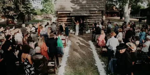 A wedding ceremony in progress with a bride and groom at the altar in front of a historic log cabin, surrounded by seated guests.