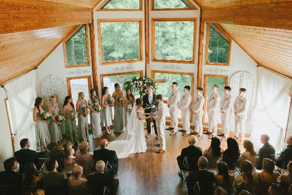 A wedding ceremony in progress inside a wooden hall with large windows, featuring a bride, groom, and wedding party.