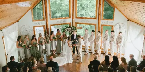 A wedding ceremony in progress inside a wooden hall with large windows, featuring a bride, groom, and wedding party.