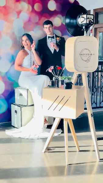 A bride and groom posing with props next to a handcrafted wooden photo booth station featuring the Bokeh & Birch logo.