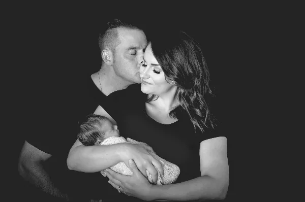 A black-and-white studio photograph of a man kissing a woman on the forehead while she holds a sleeping newborn baby.