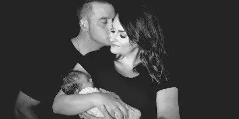 A black-and-white studio photograph of a man kissing a woman on the forehead while she holds a sleeping newborn baby.
