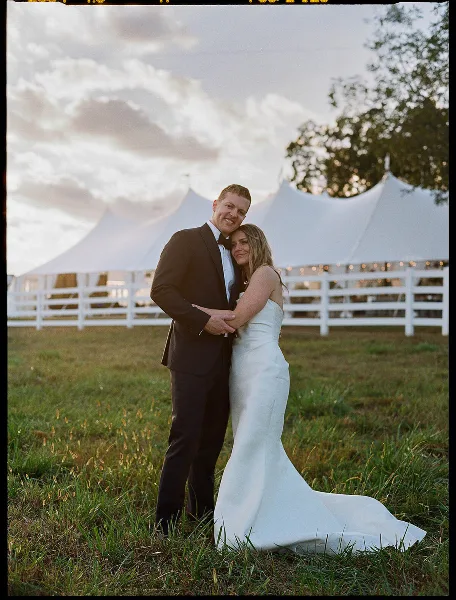A bride and groom embracing in a grassy field with a white event tent and white fence in the background.