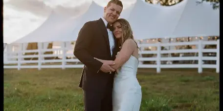 A bride and groom embracing in a grassy field with a white event tent and white fence in the background.