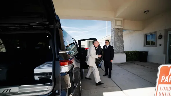 A professional chauffeur in a suit opens the door of a black luxury SUV for a passenger at a hotel entrance.