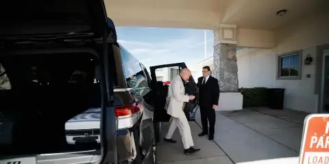 A professional chauffeur in a suit opens the door of a black luxury SUV for a passenger at a hotel entrance.