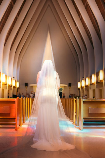 A bride in a white wedding dress and veil standing in the aisle of a church with arched ceilings and wooden pews.
