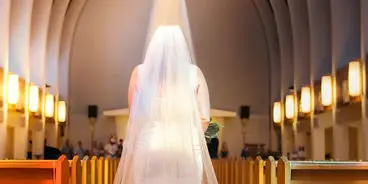 A bride in a white wedding dress and veil standing in the aisle of a church with arched ceilings and wooden pews.