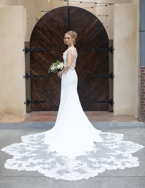 A bride in a white lace wedding dress holding a bouquet, standing in front of a large wooden arched door.