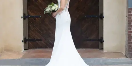 A bride in a white lace wedding dress holding a bouquet, standing in front of a large wooden arched door.
