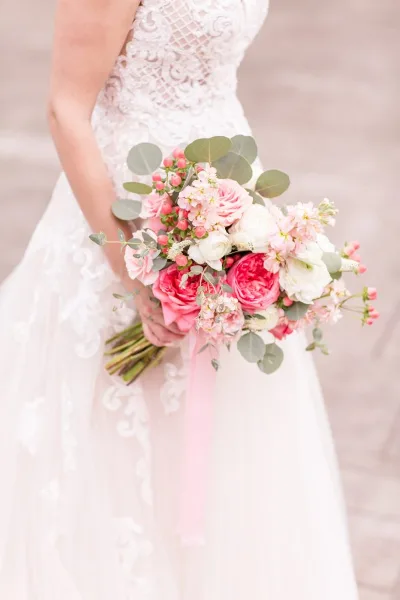 A bride in a white lace wedding dress holding a bouquet of pink and white roses with eucalyptus leaves.
