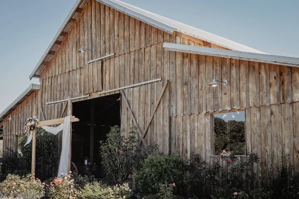 Exterior view of a rustic wooden barn wedding venue with a decorated archway and surrounding greenery.