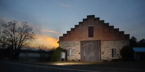 Exterior view of a historic stone and brick barn with a stepped gable roof at dusk.