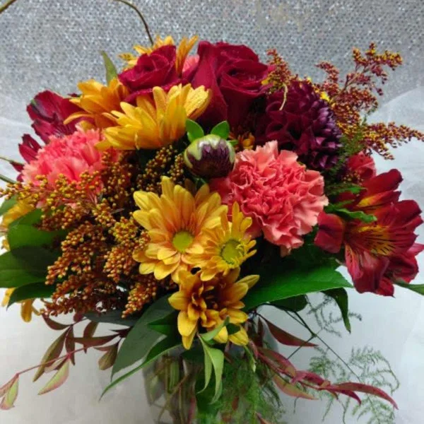 A vibrant mixed bouquet featuring red roses, yellow daisies, and pink carnations arranged in a clear glass vase.