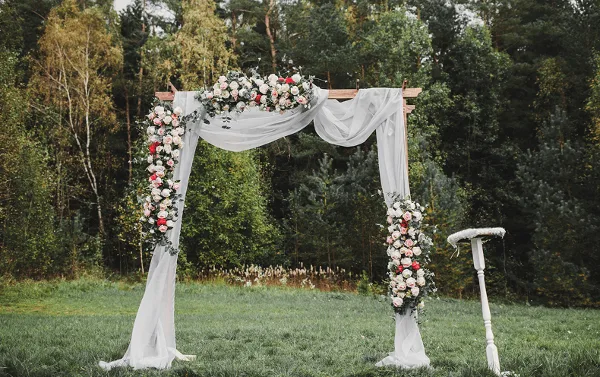 Outdoor wedding archway decorated with flowers and white drapery, with a sign stand nearby.