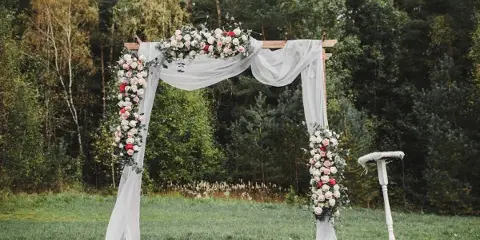 Outdoor wedding archway decorated with flowers and white drapery, with a sign stand nearby.