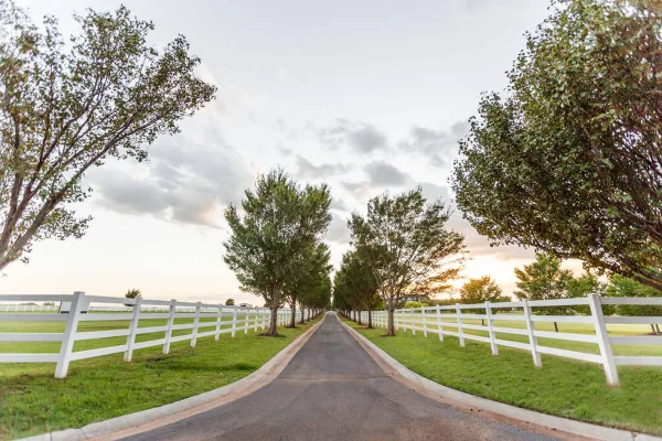 A paved tree-lined road flanked by white fences and manicured grass at a private estate venue.