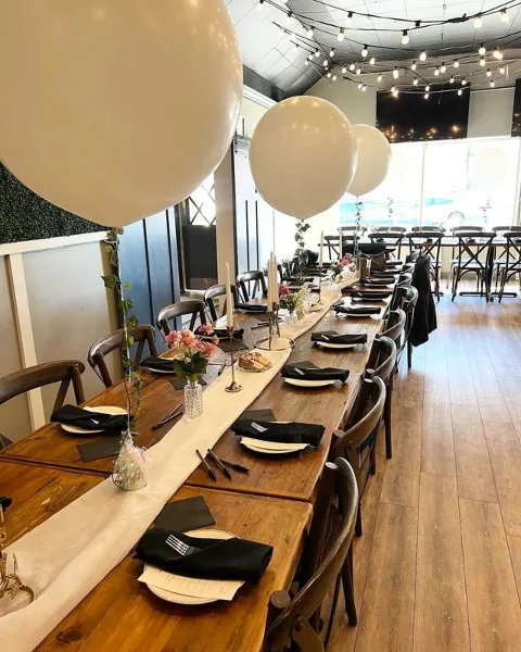 Long wooden table set for a meal in a restaurant with white balloons and string lights.