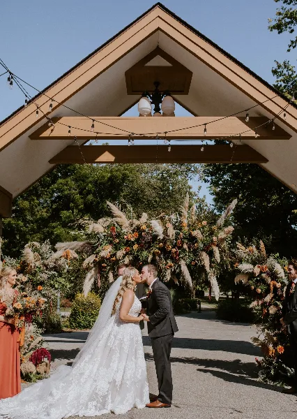 A bride and groom kissing under a wooden wedding arch decorated with string lights and a large floral arrangement at an outdoor venue.