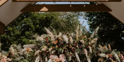 A bride and groom kissing under a wooden wedding arch decorated with string lights and a large floral arrangement at an outdoor venue.