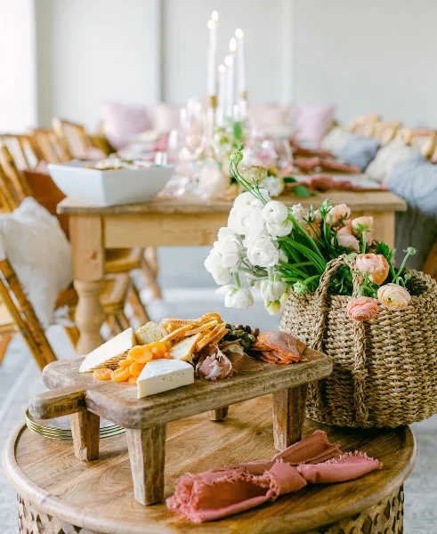 A rustic wooden board holding an assortment of cheeses, cured meats, dried fruit, and bread, placed next to a woven basket of white and pink flowers on a wooden table.