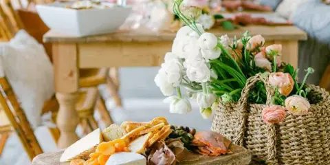 A rustic wooden board holding an assortment of cheeses, cured meats, dried fruit, and bread, placed next to a woven basket of white and pink flowers on a wooden table.