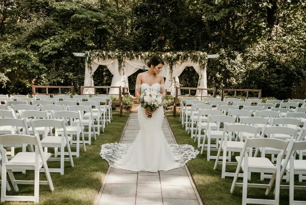 Bride in a white gown holding a bouquet standing in an outdoor garden ceremony aisle with white chairs and a floral arch.