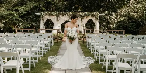 Bride in a white gown holding a bouquet standing in an outdoor garden ceremony aisle with white chairs and a floral arch.