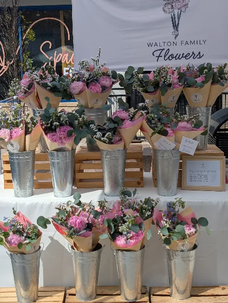 Rows of wrapped pink and white flower bouquets in metal buckets at a market table with a Walton Family Flowers banner.