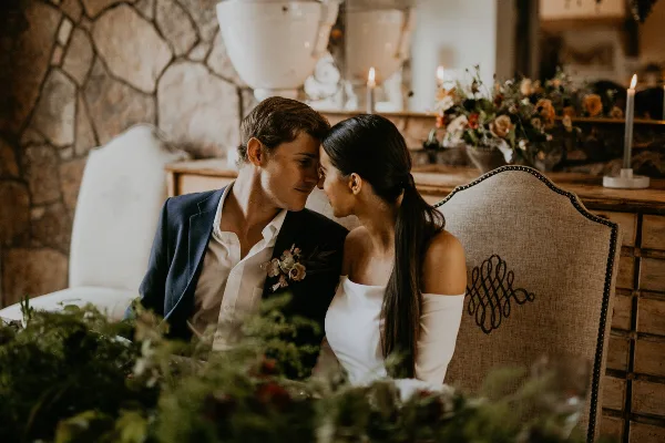 A bride and groom in formal wedding attire sitting closely together at a decorated table with floral arrangements and candles.