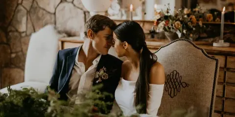 A bride and groom in formal wedding attire sitting closely together at a decorated table with floral arrangements and candles.
