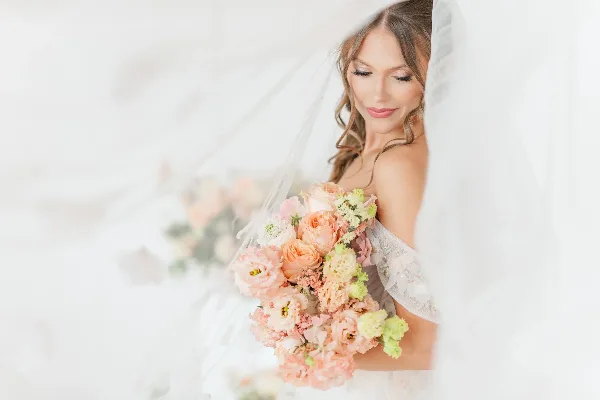 A bride in a white gown and veil holding a bouquet of pink, peach, and cream flowers.