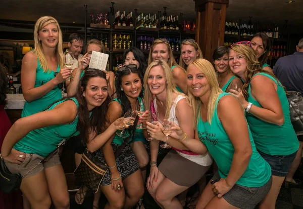 A group of smiling women in matching teal shirts posing with wine glasses at a bar.