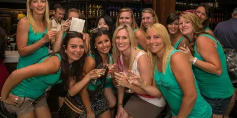 A group of smiling women in matching teal shirts posing with wine glasses at a bar.