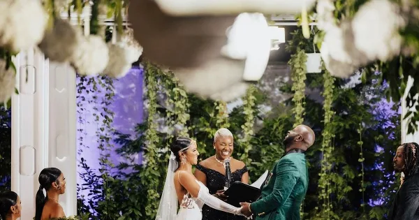 A wedding officiant with short white hair stands at the altar between a bride and groom during a ceremony, with a floral backdrop.