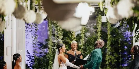 A wedding officiant with short white hair stands at the altar between a bride and groom during a ceremony, with a floral backdrop.