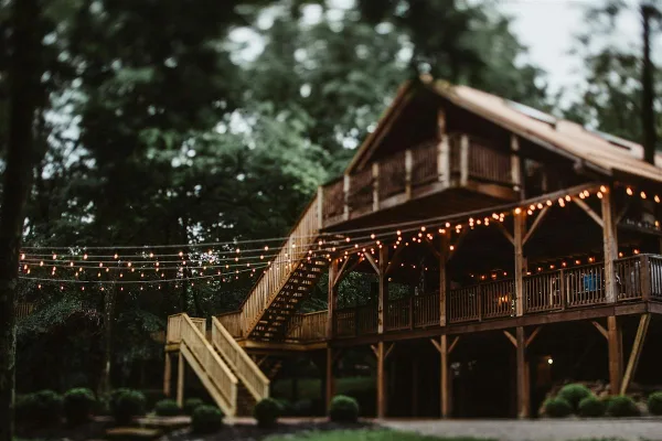 A large wooden venue building with a covered porch and string lights, surrounded by trees.