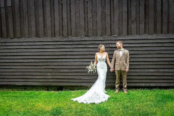A bride in a white lace gown and a groom in a tan suit standing hand-in-hand on a lawn in front of a dark wooden barn wall.