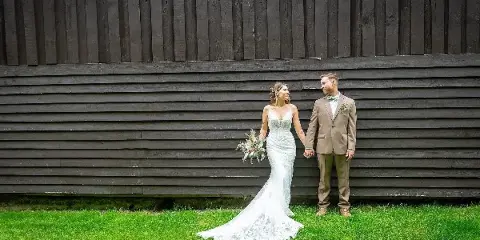 A bride in a white lace gown and a groom in a tan suit standing hand-in-hand on a lawn in front of a dark wooden barn wall.