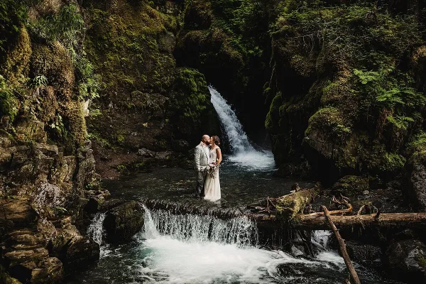 A bride and groom in wedding attire standing together in front of a forest waterfall.